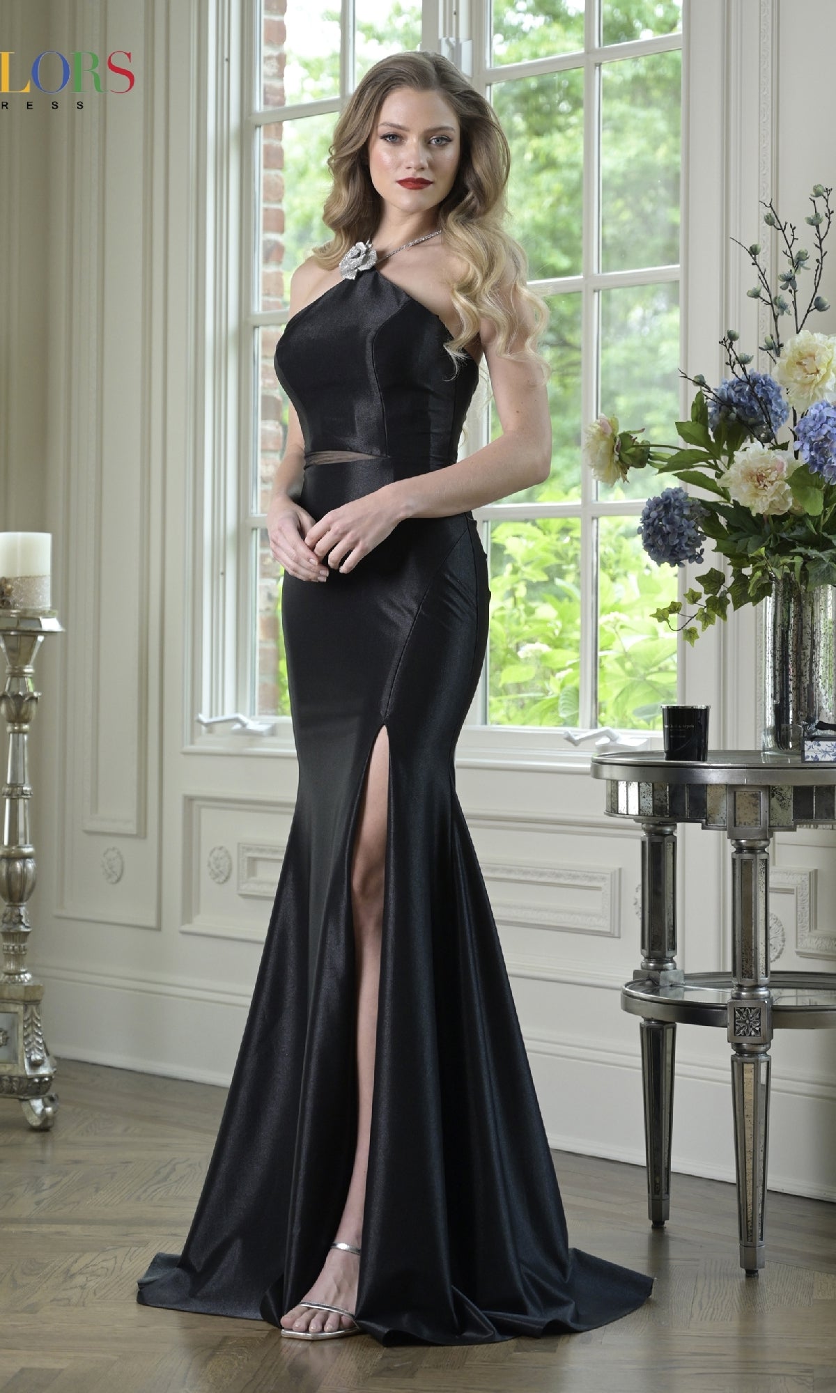 A woman stands indoors near a window and a table with flowers, wearing the Colors Dress 3926 Cut-Out Sleek Long Prom Dress in black, featuring a corset-style bodice and thigh-high slit.